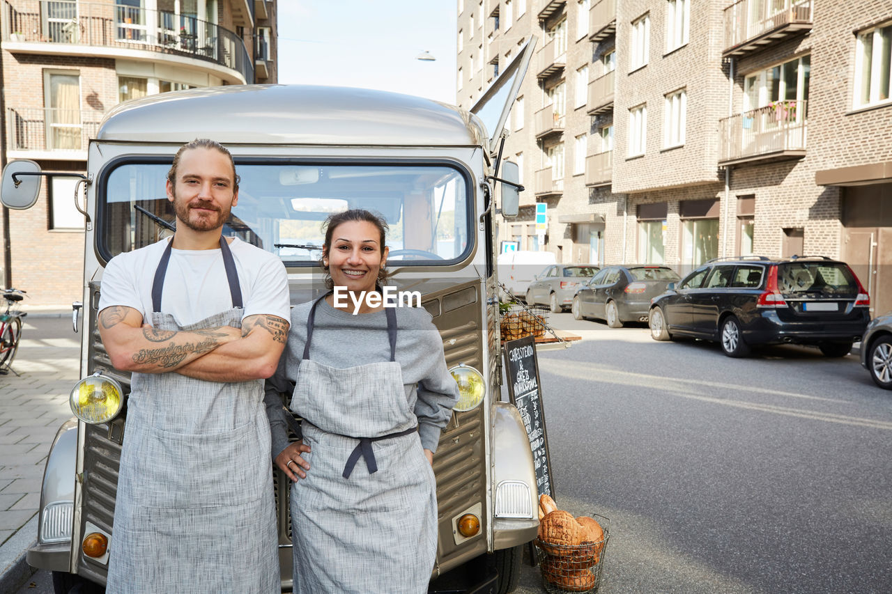 Portrait of confident male and female owners standing in front of food truck parked on city street