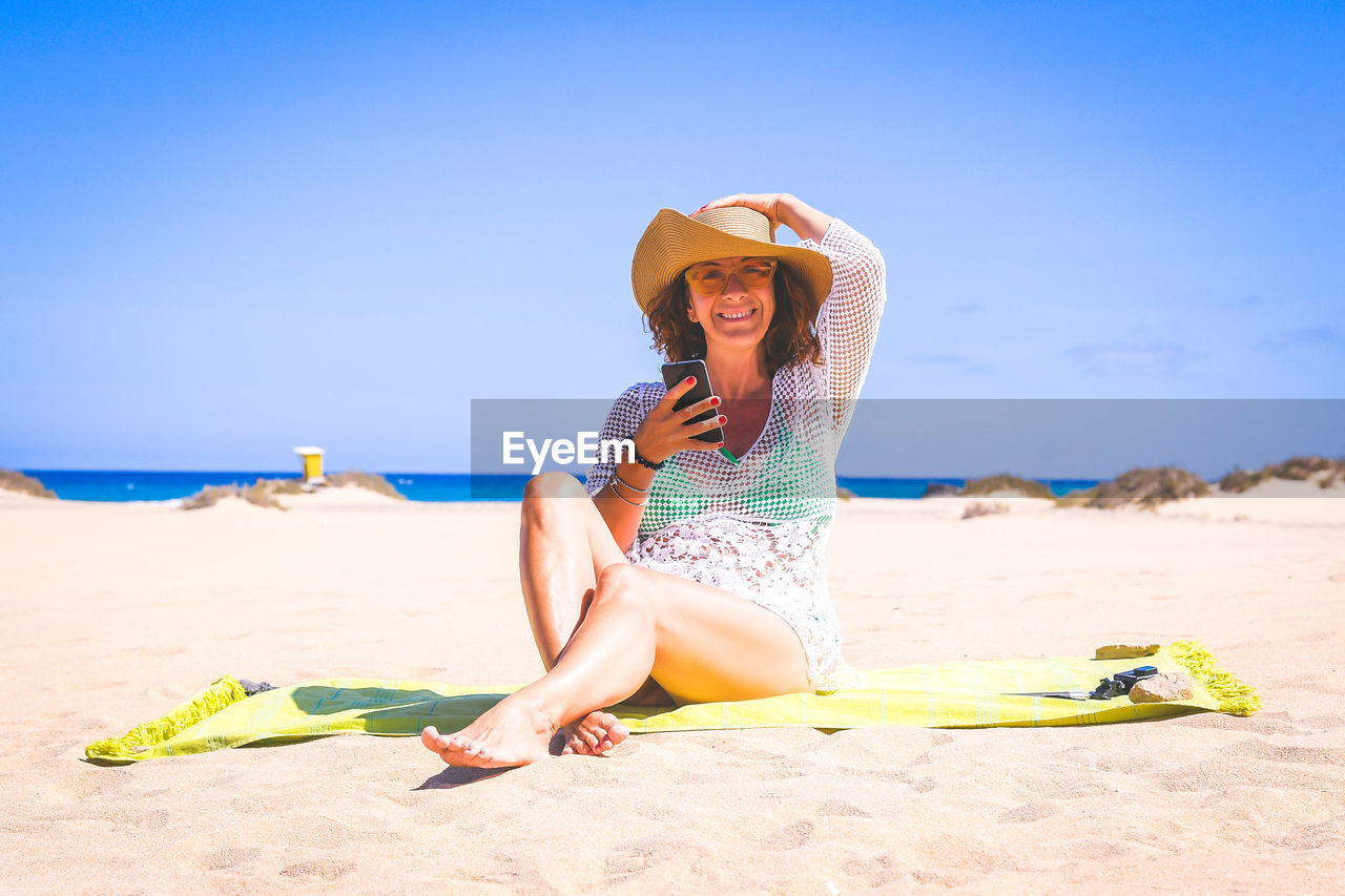Portrait of smiling mature woman using smart phone while sitting at beach against sky during sunny day