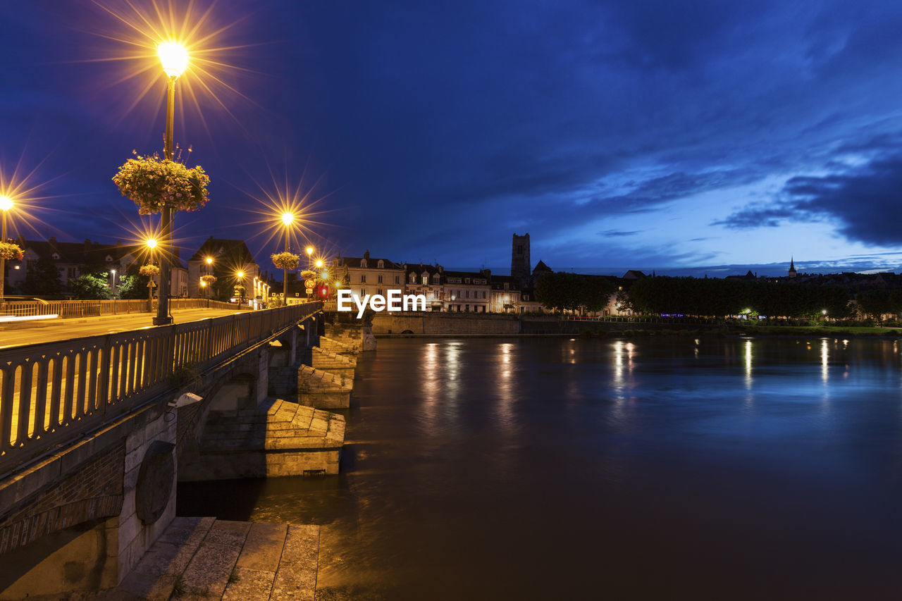 illuminated bridge over river against cloudy sky at night