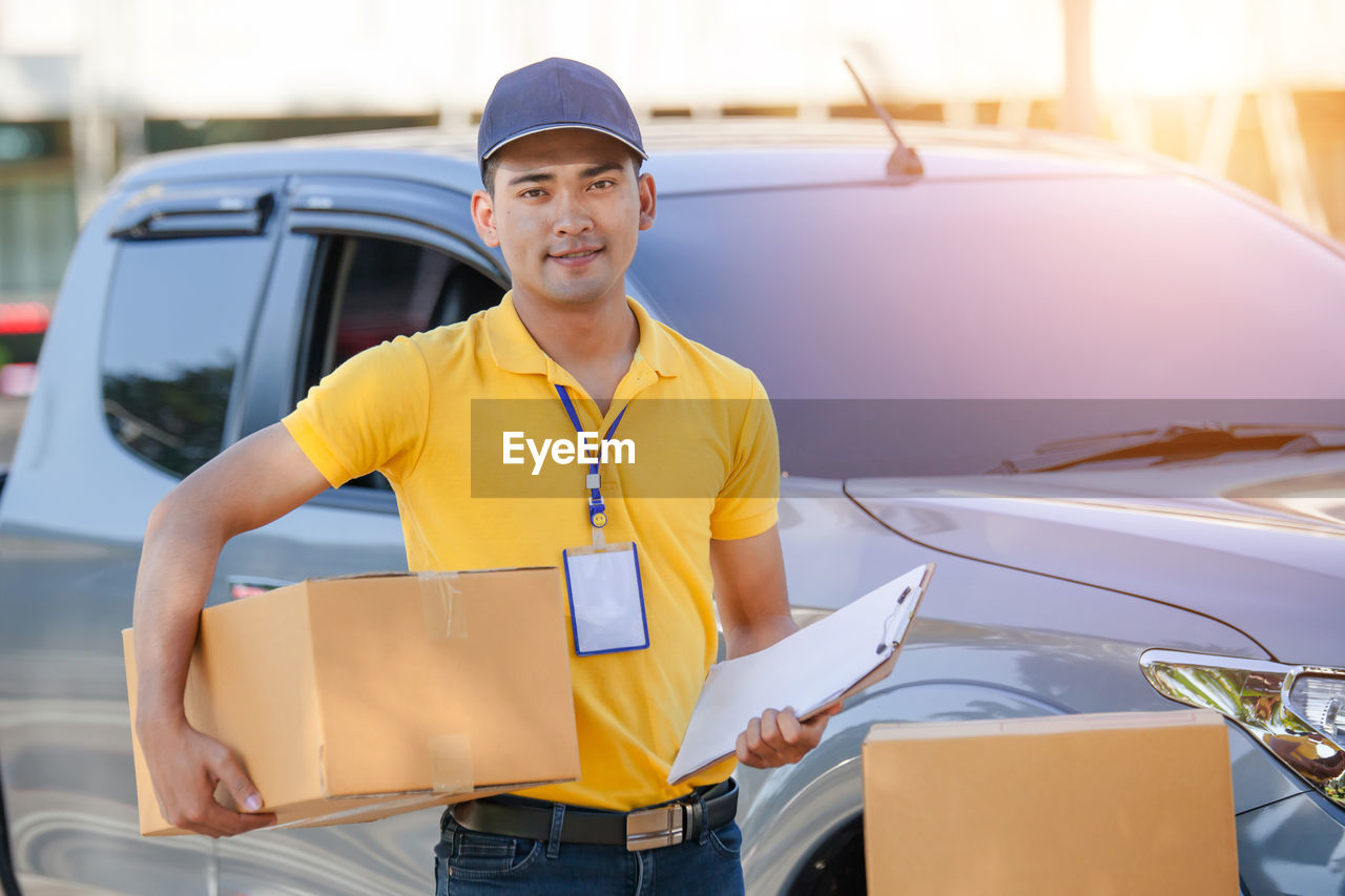 Portrait of smiling delivery person with box standing against car