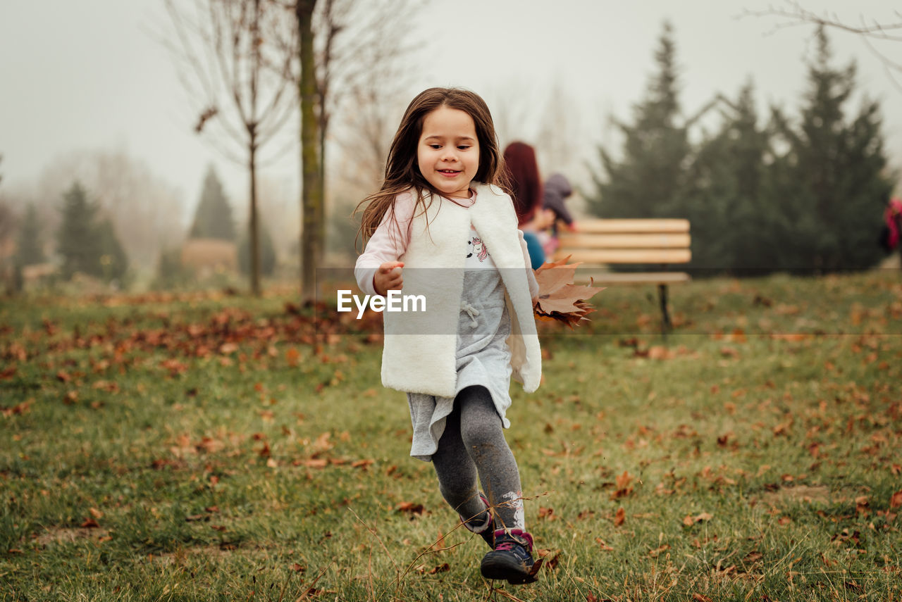 Little girl running away from mom and little sister that stay in the background on a bench