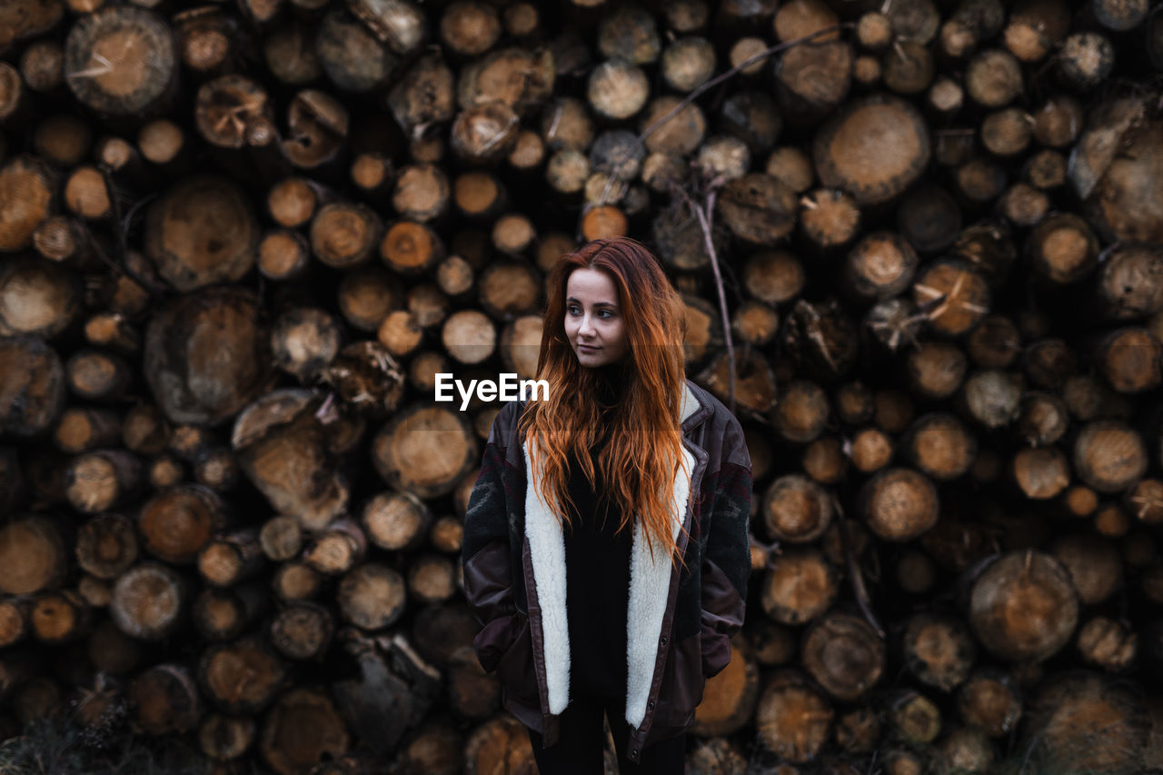 Young red haired female in warm jacket standing against stacked wooden logs in autumn day in countryside