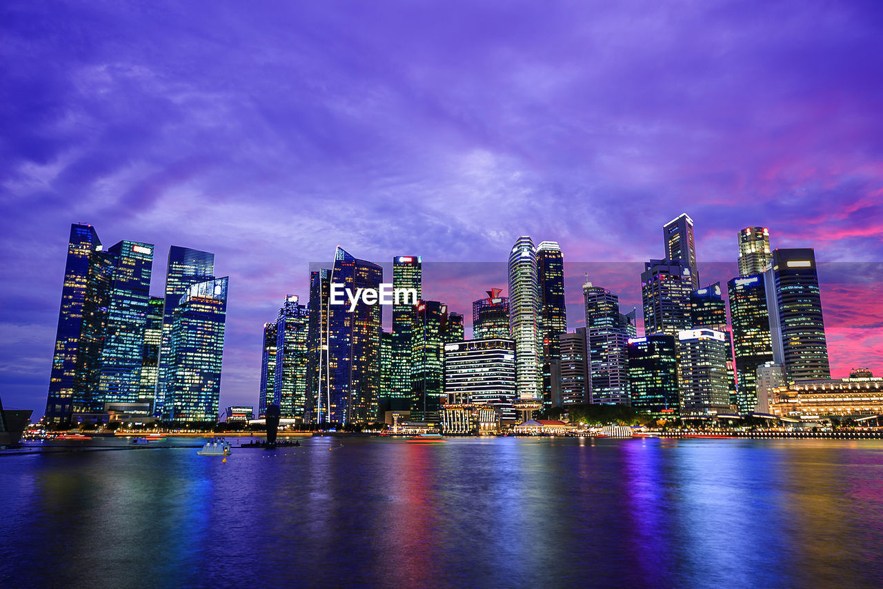 Illuminated city at waterfront against cloudy sky