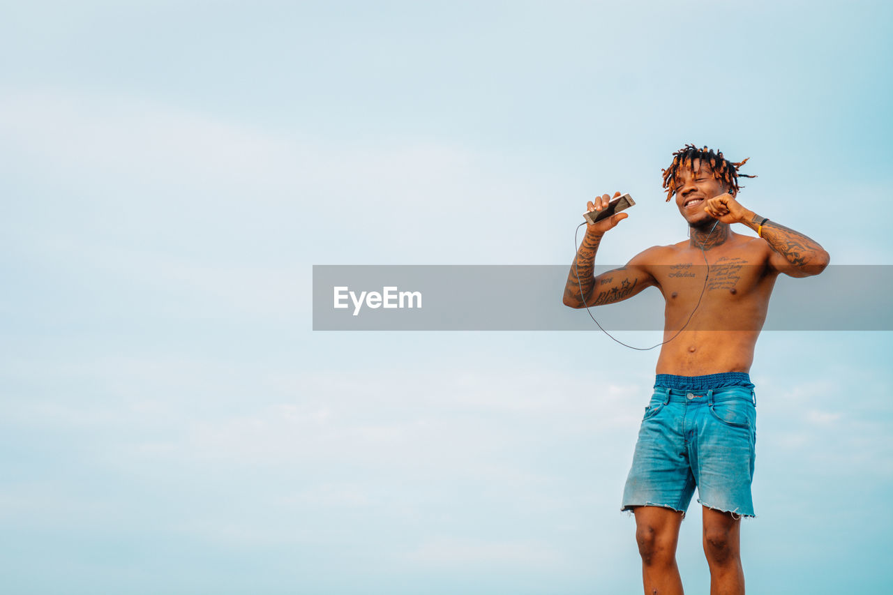 Full length of shirtless man listening to music against sky