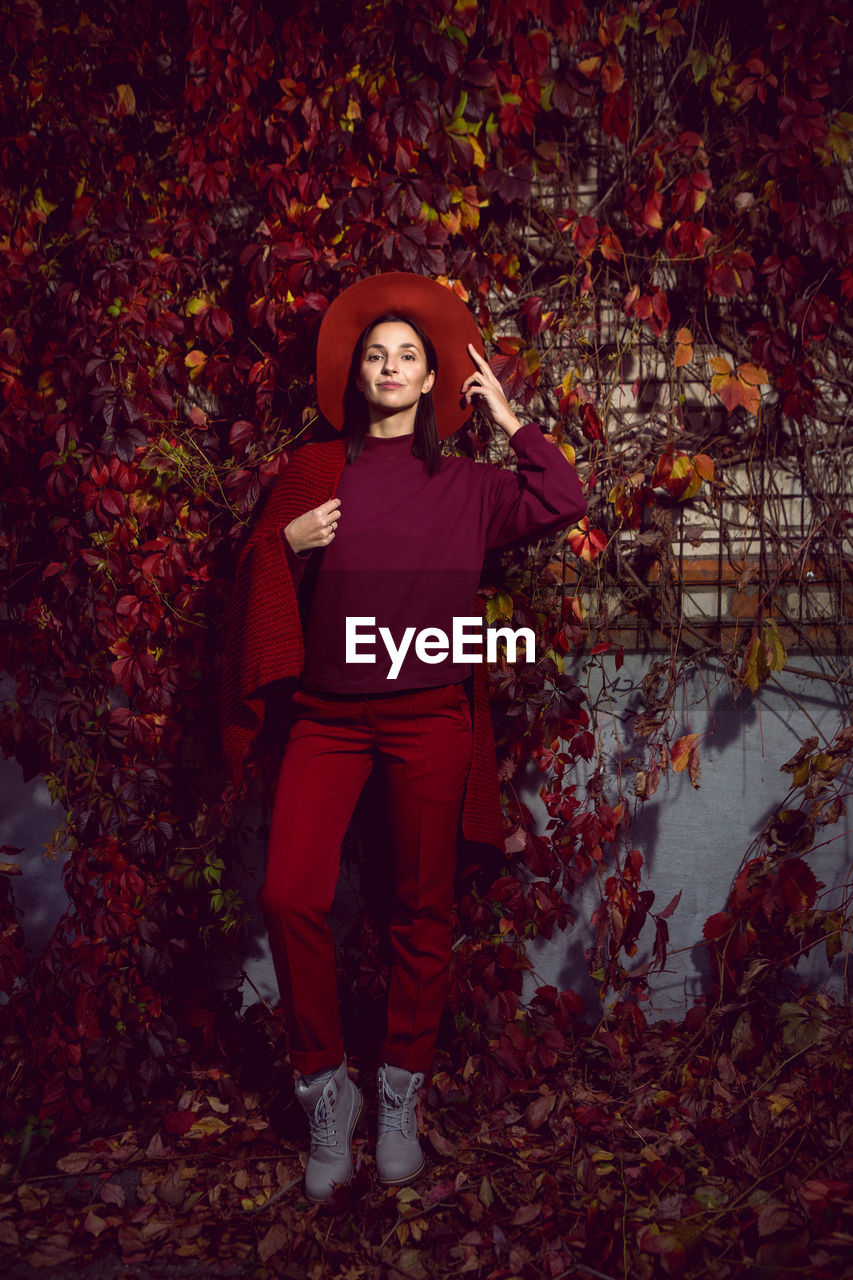 Woman in a red hat and red sweater stands on a background of orange leaves grapes wall autumn