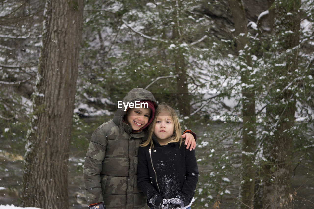 Portrait of happy brother standing with sister against trees during winter