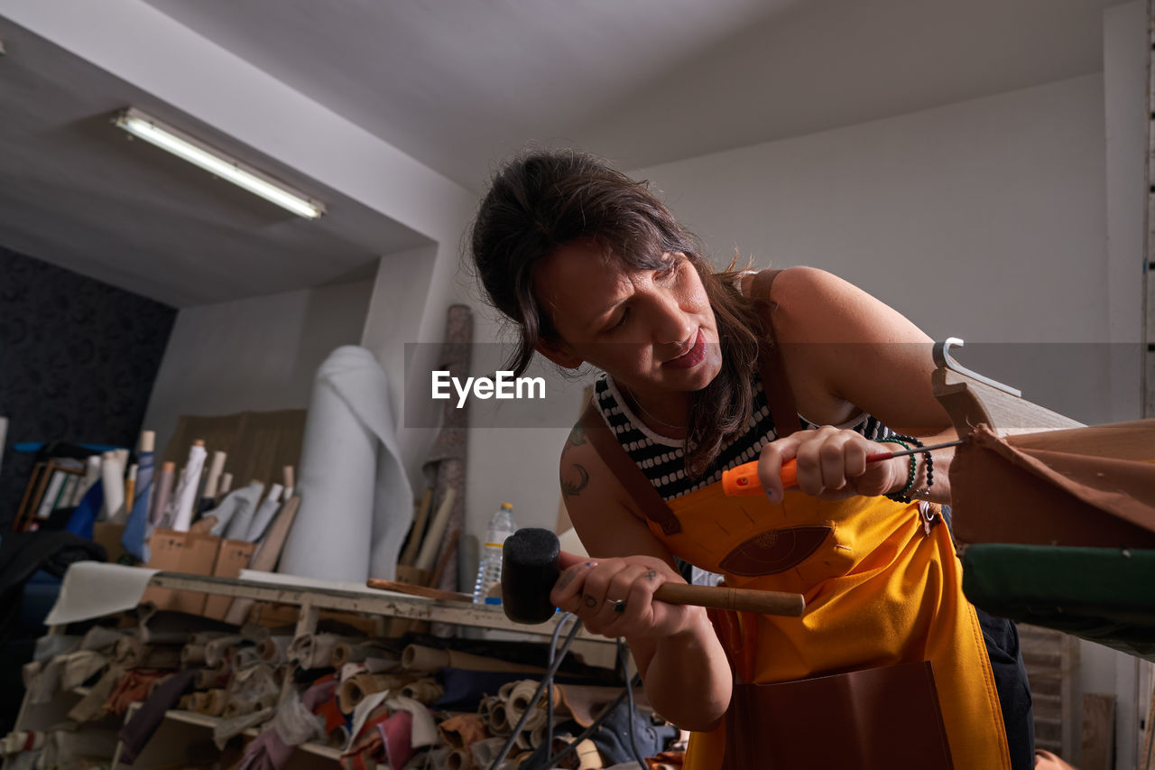 Side view of concentrated adult female master with dark hair in apron hitting on screwdriver with hammer while creating wooden furniture sitting at workbench in traditional joinery