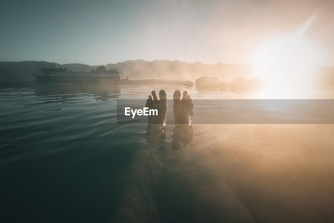 Low section of person relaxing in lake against sky during sunrise