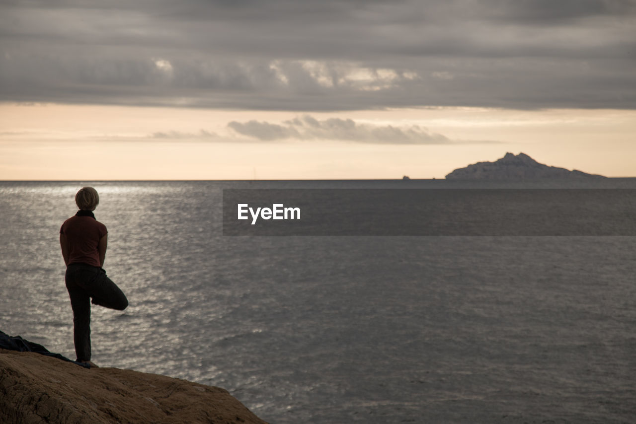 Rear view of woman practicing yoga on rock by sea during sunset