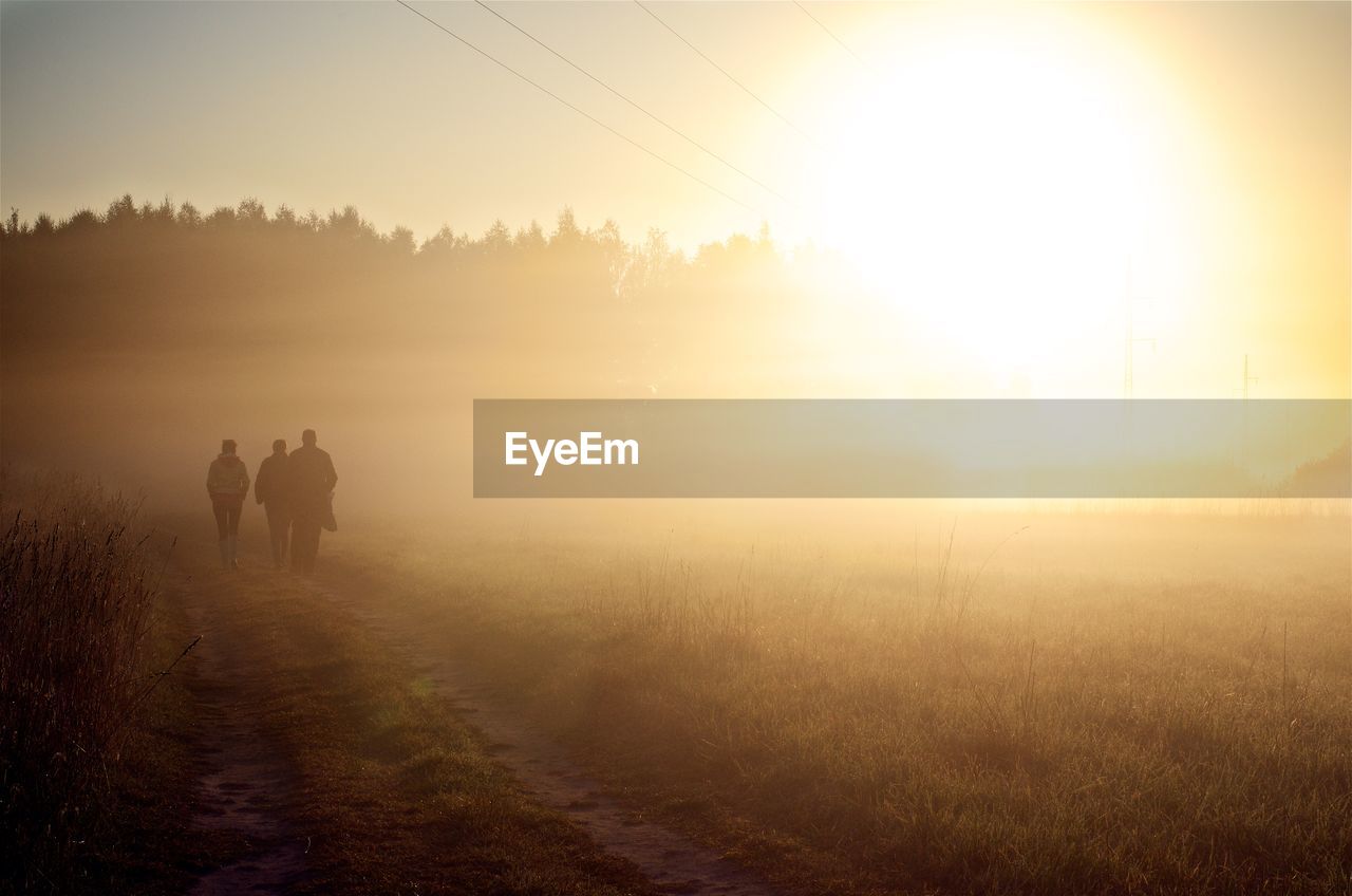 REAR VIEW OF PEOPLE WALKING ON FIELD DURING SUNSET