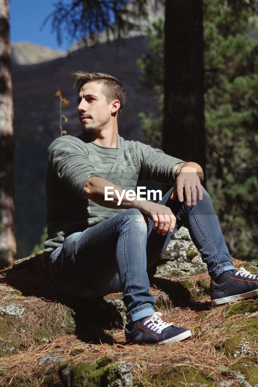 Young man looking away while sitting on rock in forest