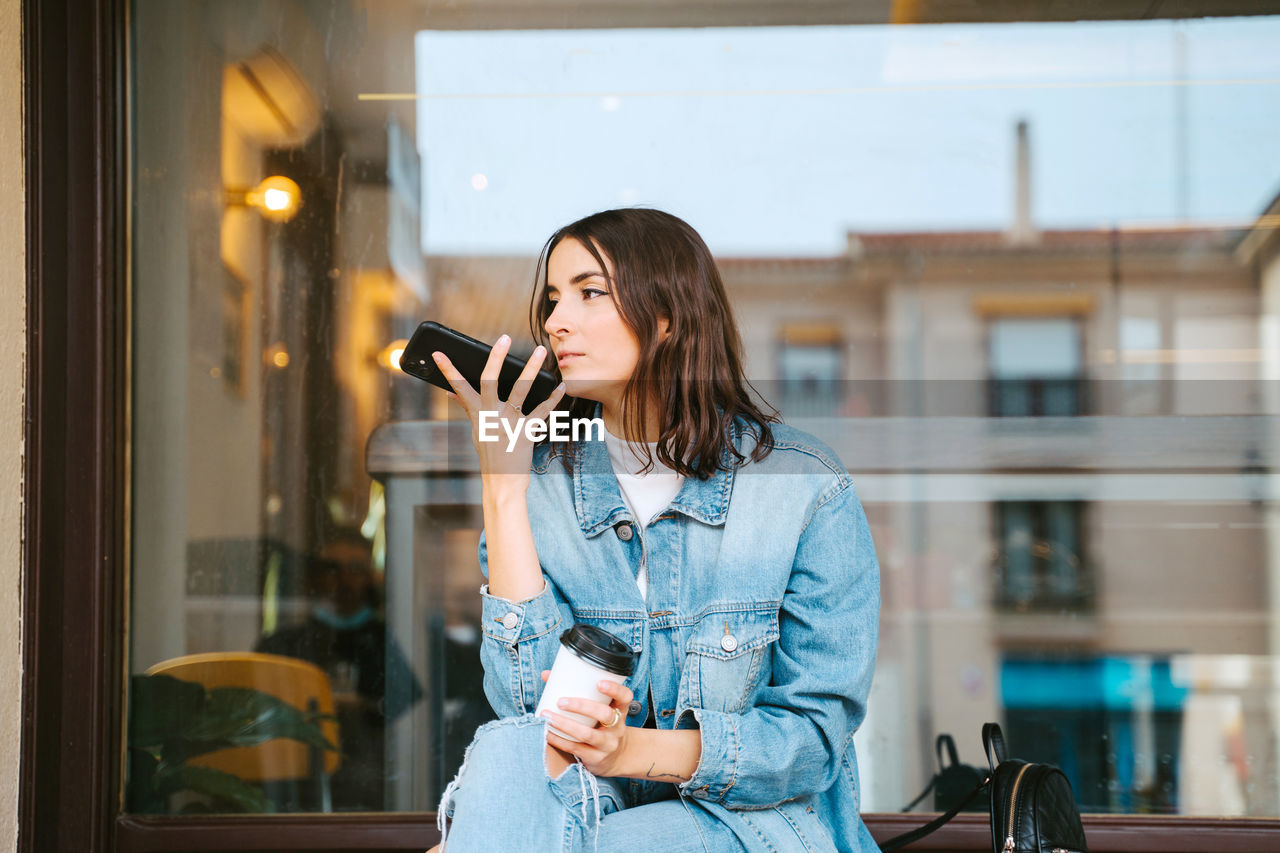 Modern young woman in trendy denim outfit with ripped jeans holding cup of takeaway coffee and speaking on mobile phone while sitting on bench near urban building