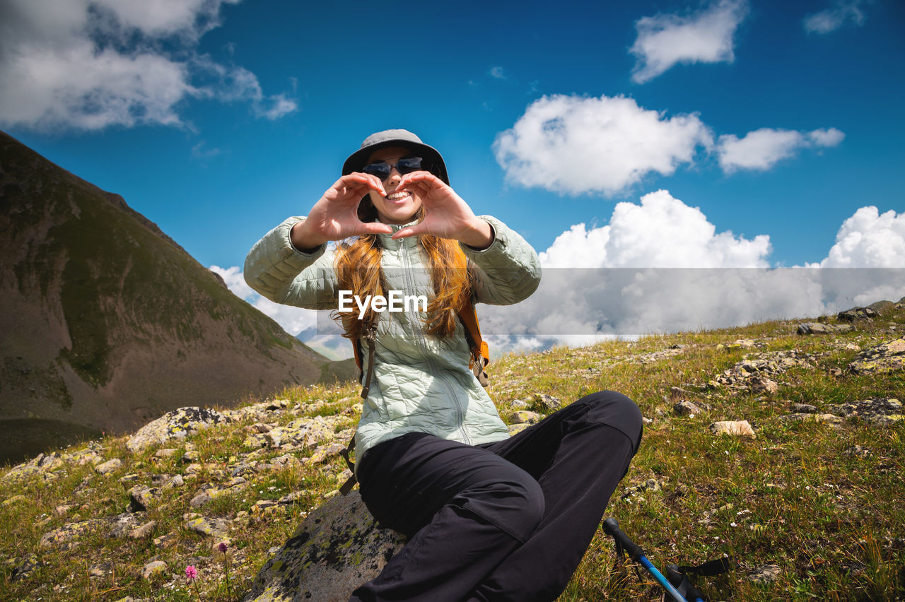 Smiling young woman showing fingers gesture sign of heart, love in mountains, happy tourist