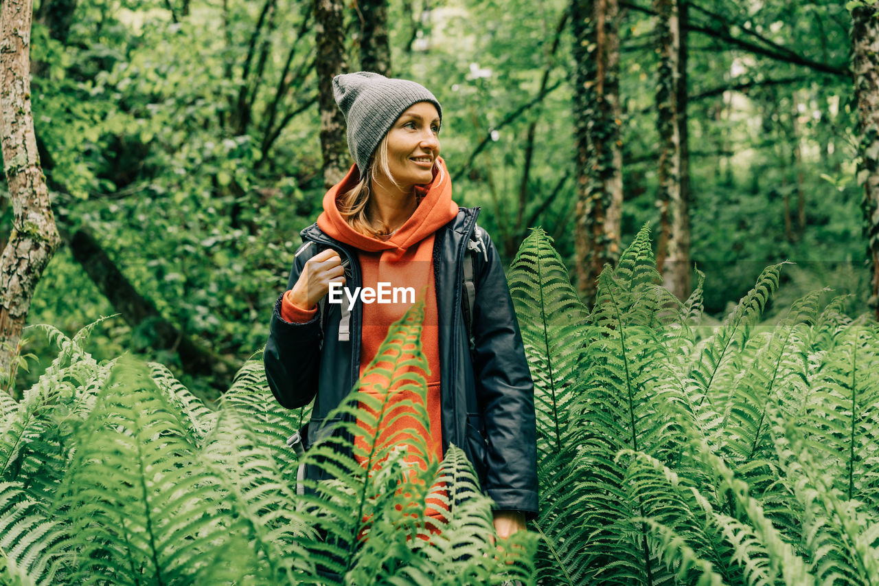 Young active woman hiker in a jacket and a hat with a backpack in a thicket of ferns in the forest