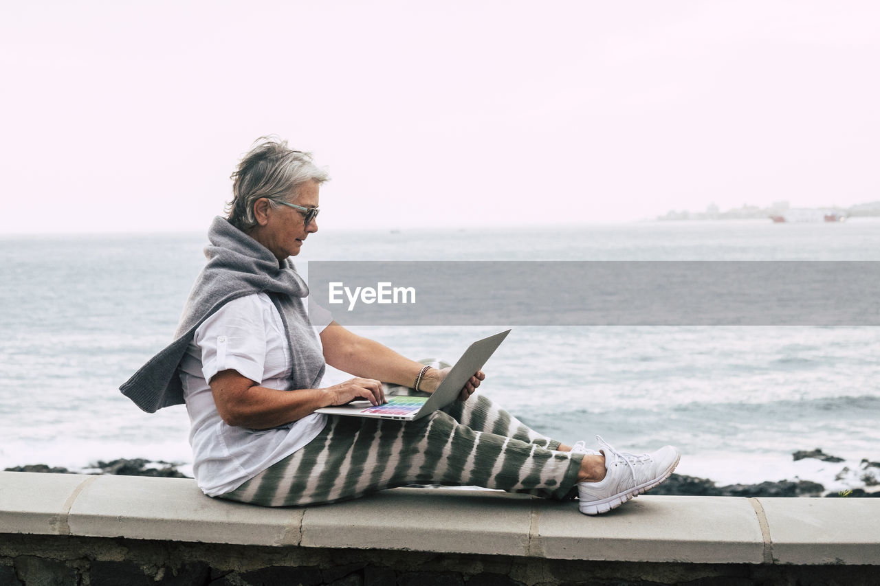 Senior woman sitting on wall by the sea, using laptop