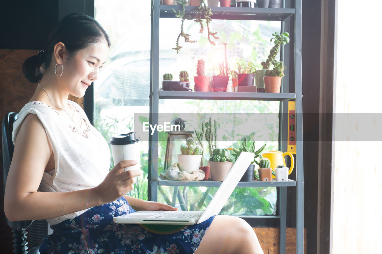 Side view of woman using laptop while sitting against potted plants on shelves at home