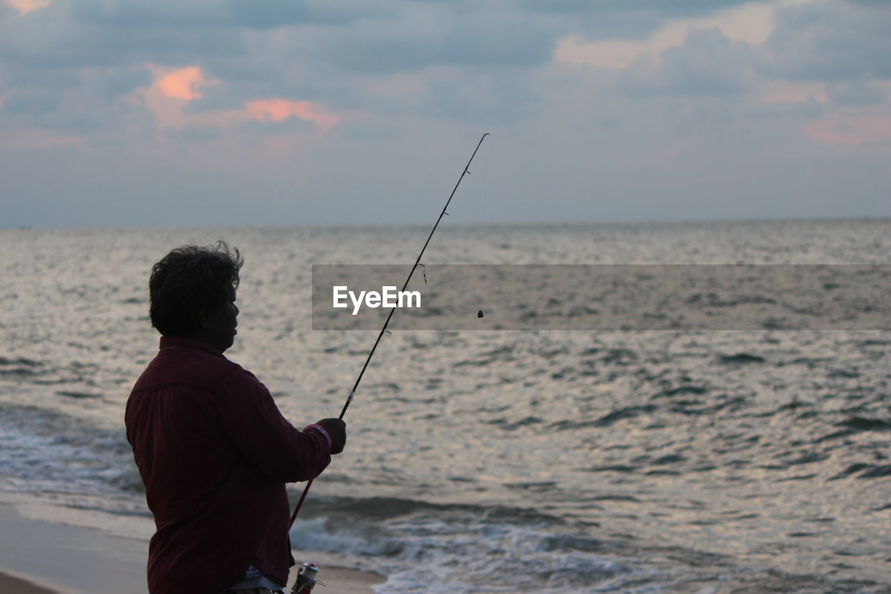 Rear view of man fishing in sea against sky during sunset