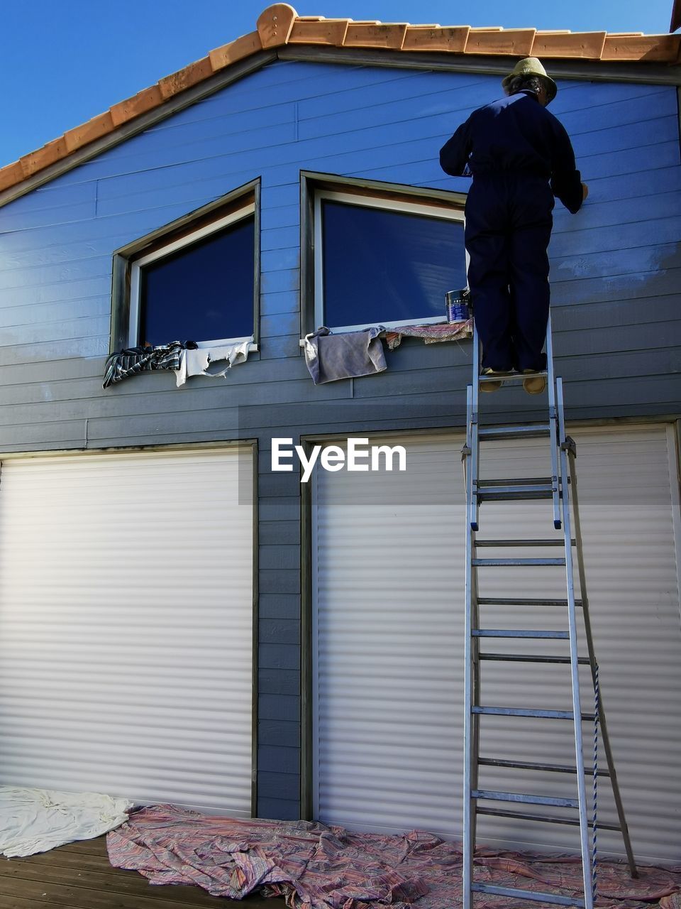 architecture, building exterior, built structure, roof, occupation, building, house, blue, working, wall, ladder, one person, men, full length, day, person, industry, home, manual worker, facade, sky, outdoors, window, adult, nature, residential district, business