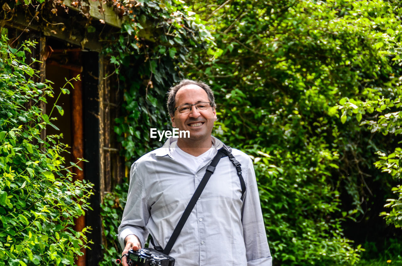 Portrait of smiling man holding camera while standing against trees