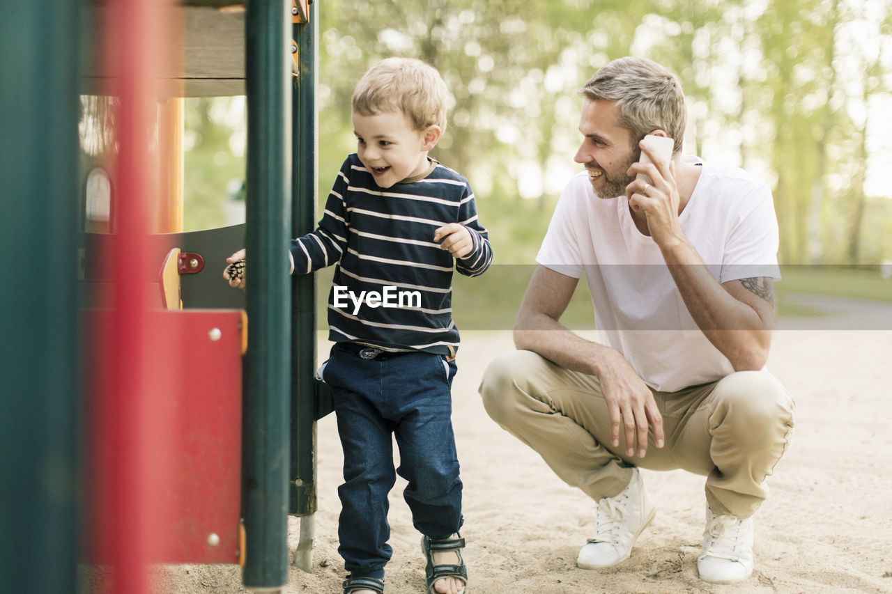 Smiling father talking on phone while looking at son playing in playground