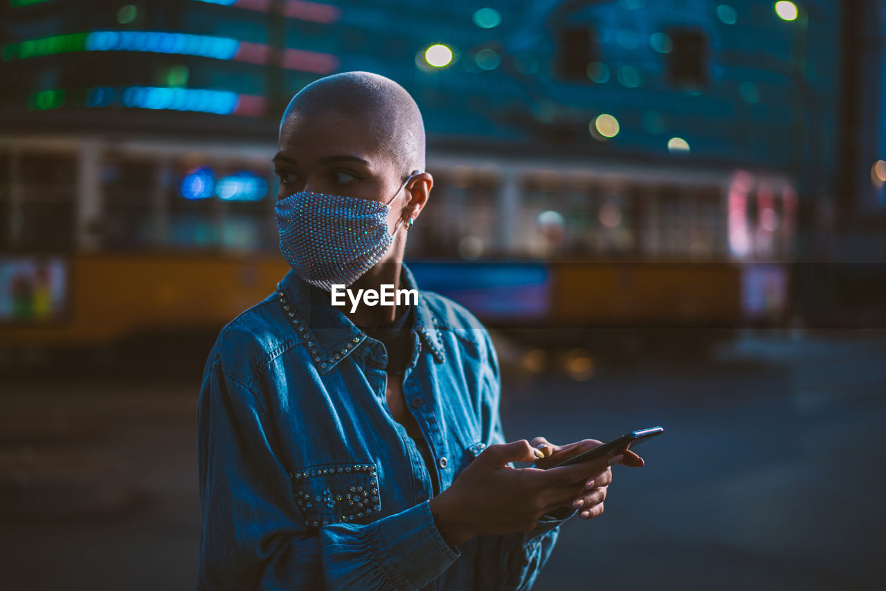 Young woman wearing mask with shaved head looking away while holding smart phone outdoors