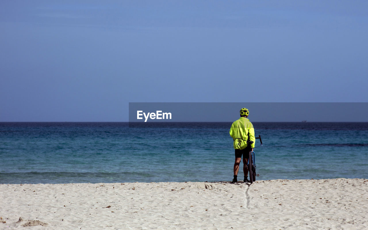 Rear view of man with bicycle standing at beach against clear sky