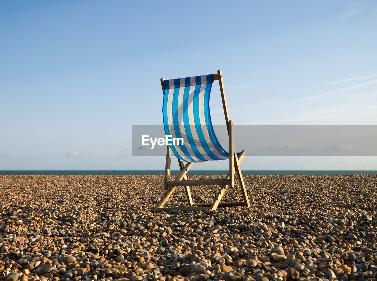 Blue and white striped empty deck chair on a pebble beach with blue sky and a sliver of sea behind