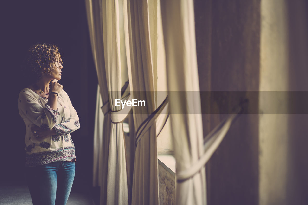 Woman looking through window while standing in corridor