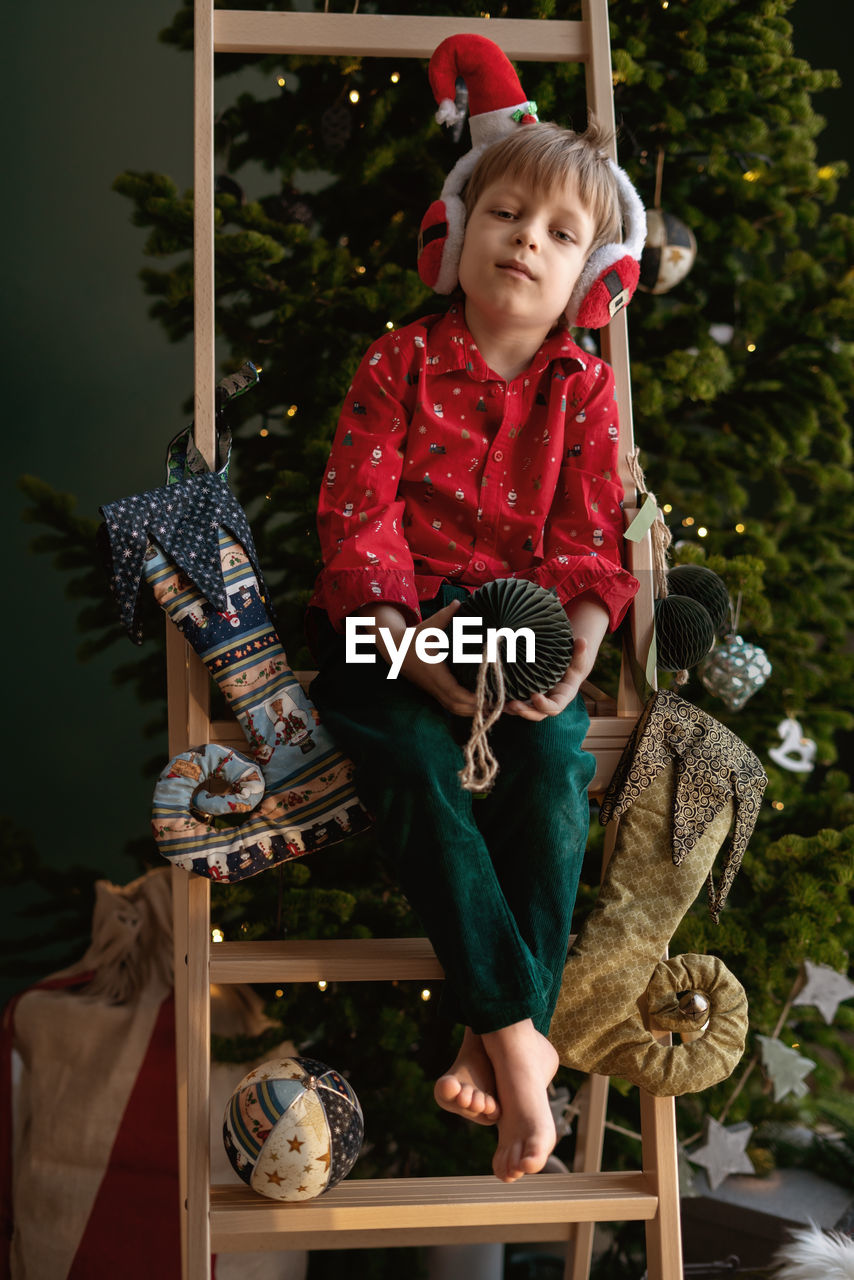 Barefoot boy in an elf costume holds craft paper deep green christmas tree ball