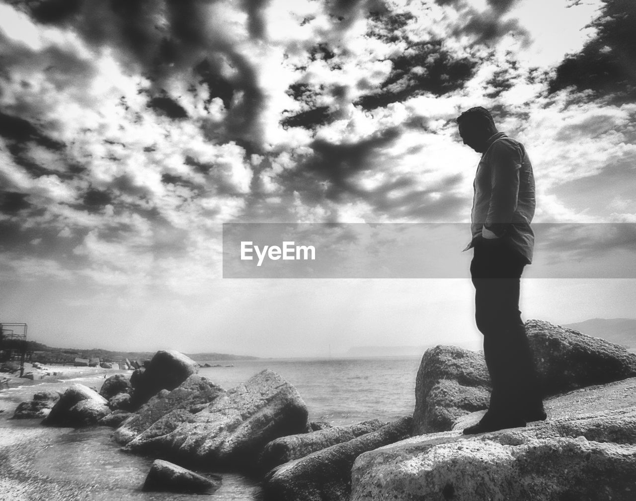 MAN STANDING ON ROCKS AT SEA SHORE AGAINST SKY