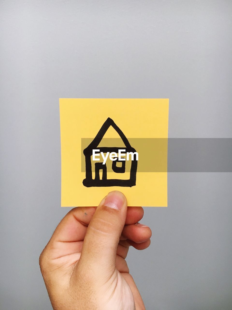 Cropped hand of person holding adhesive note with house symbol against gray background