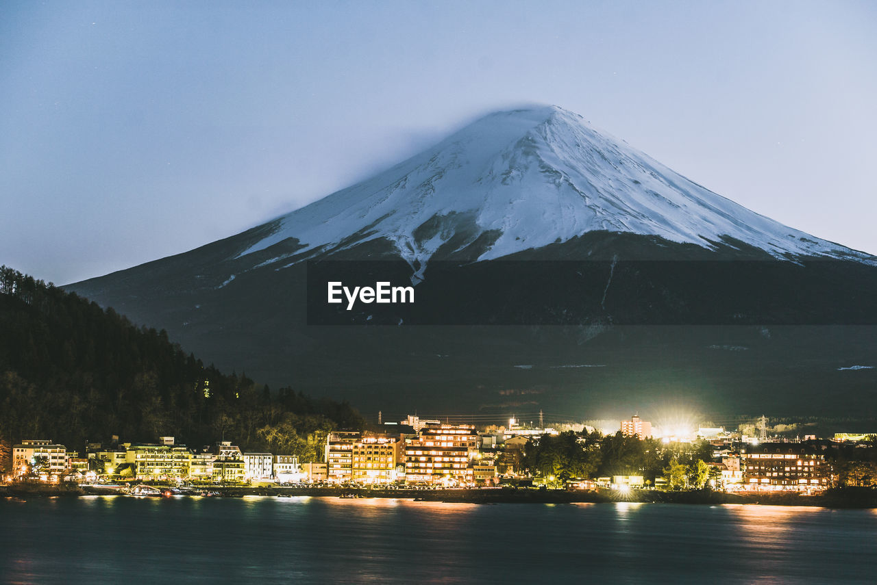 Illuminated buildings by snowcapped mountain against sky at night