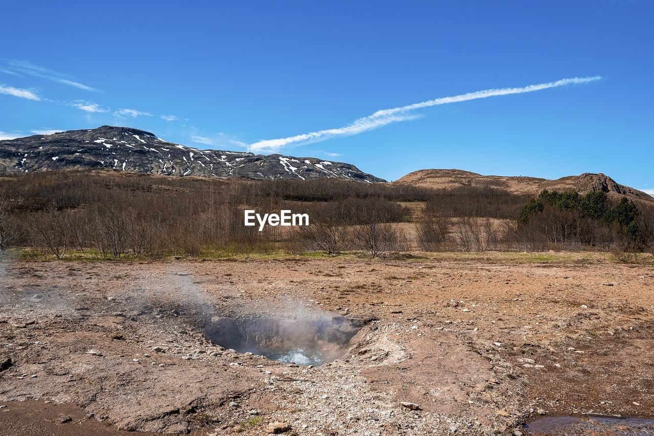 Smoke emitting from strokkur geyser amidst landscape against blue sky