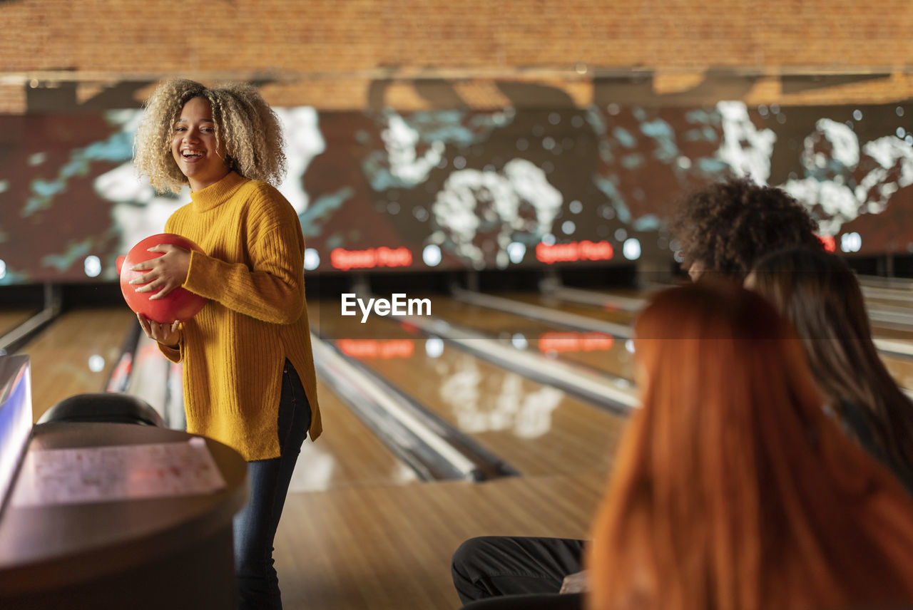 Smiling young woman holding ball with friends sitting at bowling alley