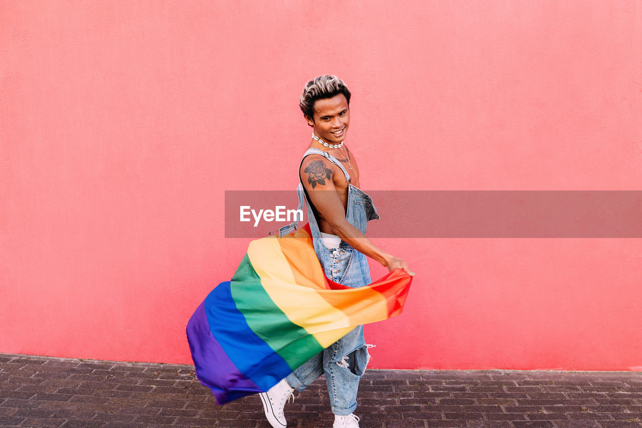 Portrait of young man holding rainbow flag in front of pink wall