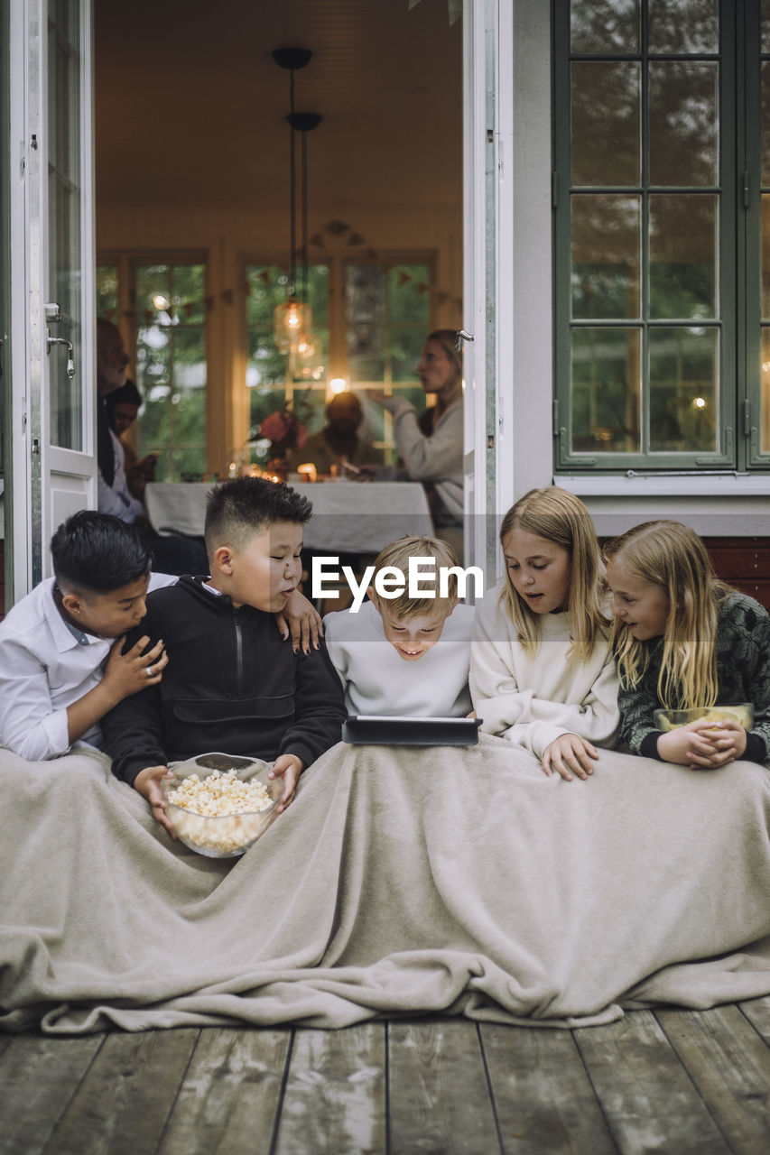 Excited friends with blanket and popcorn watching tablet pc while sitting on porch