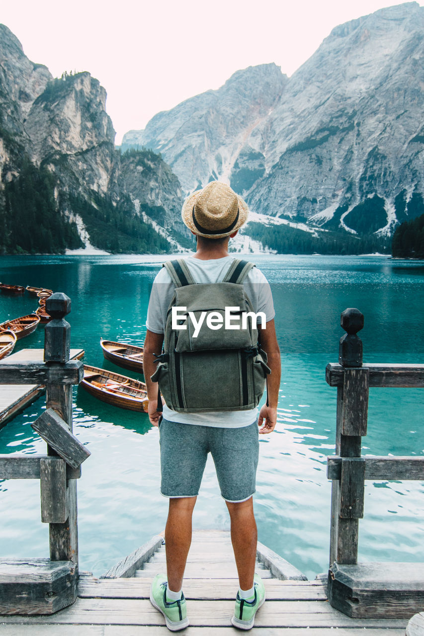 Back view of unrecognizable male traveler standing on wooden quay and admiring amazing scenery of lake with turquoise water in mountains on foggy day