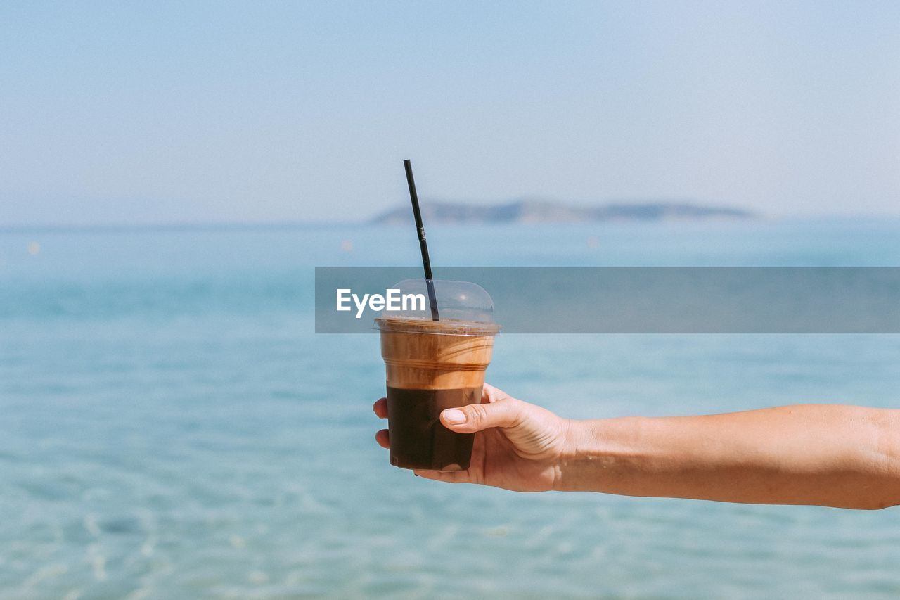 Cropped hand holding coffee against sea