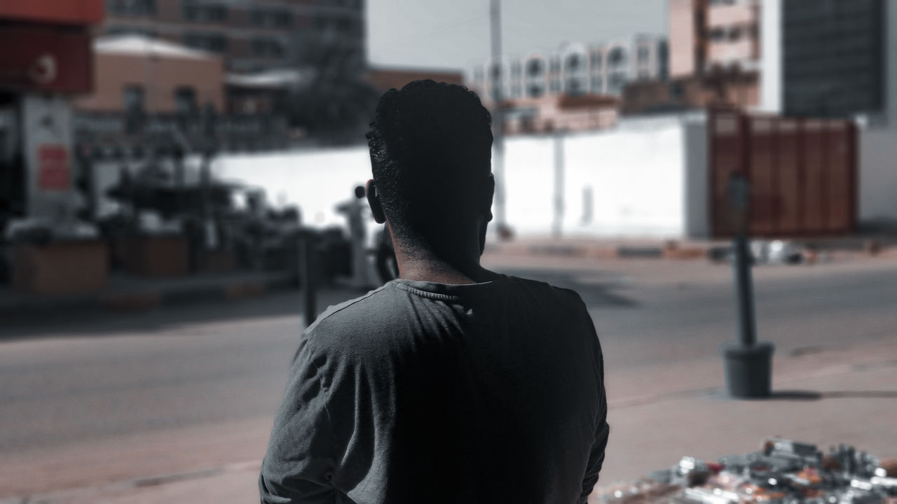 one person, rear view, city, architecture, black, adult, focus on foreground, street, standing, waist up, building exterior, day, lifestyles, men, young adult, city life, road, casual clothing, built structure, black hair, outdoors