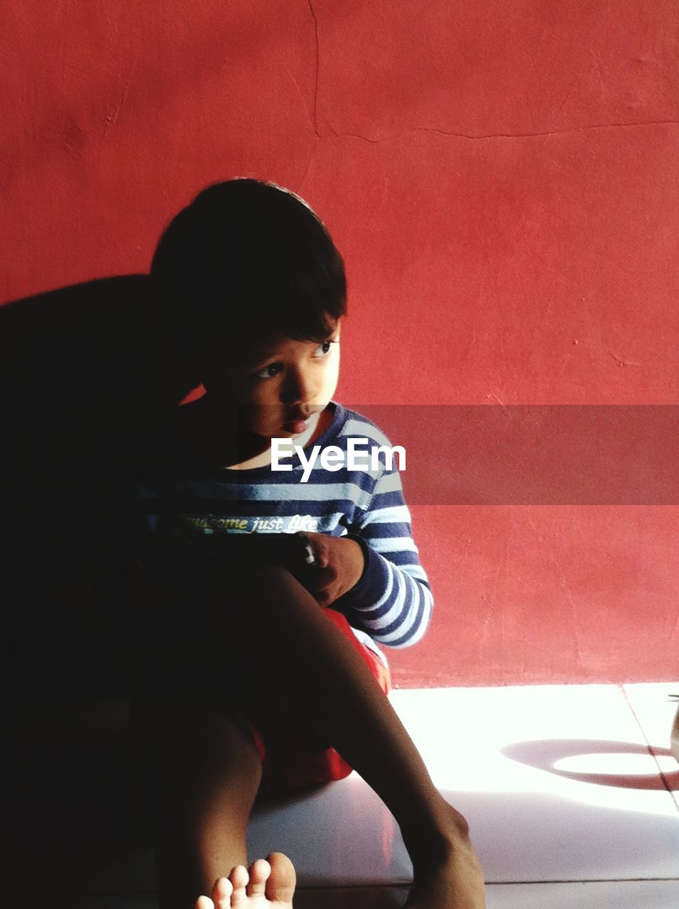 Cute boy looking away while sitting on tiled floor against wall at home