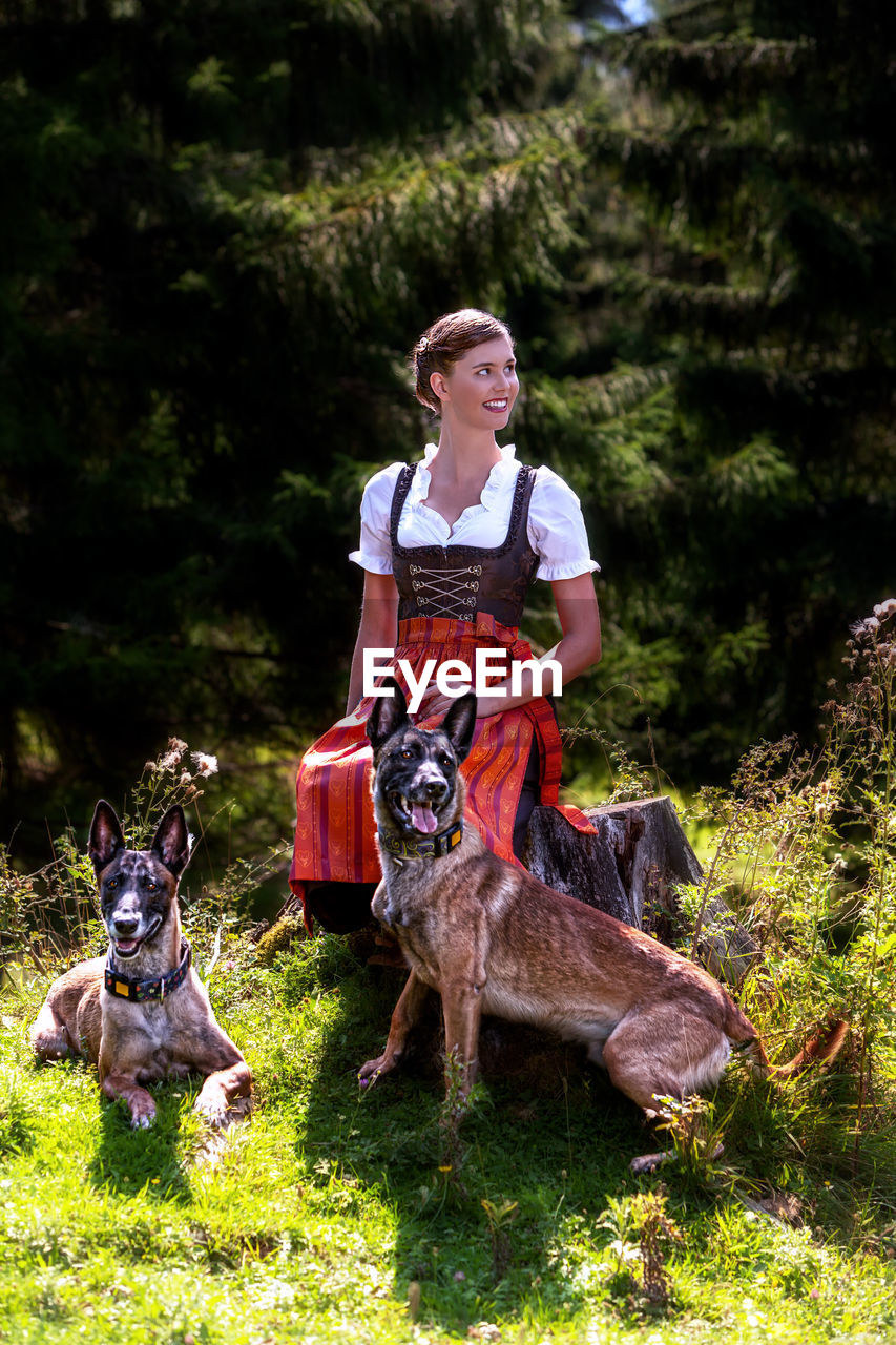 Full length of young woman with dogs sitting against trees in forest