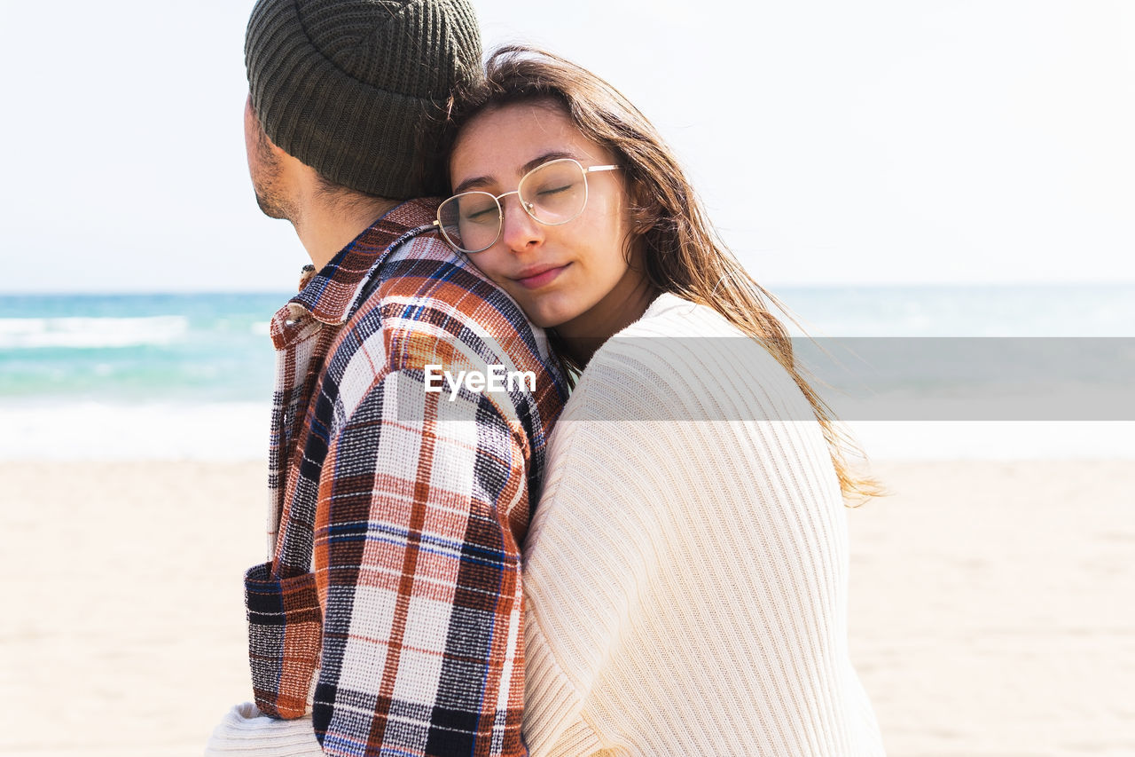 Romantic couple in casual wear cuddling while standing together on sandy beach near waving sea on sunny day in coastal area