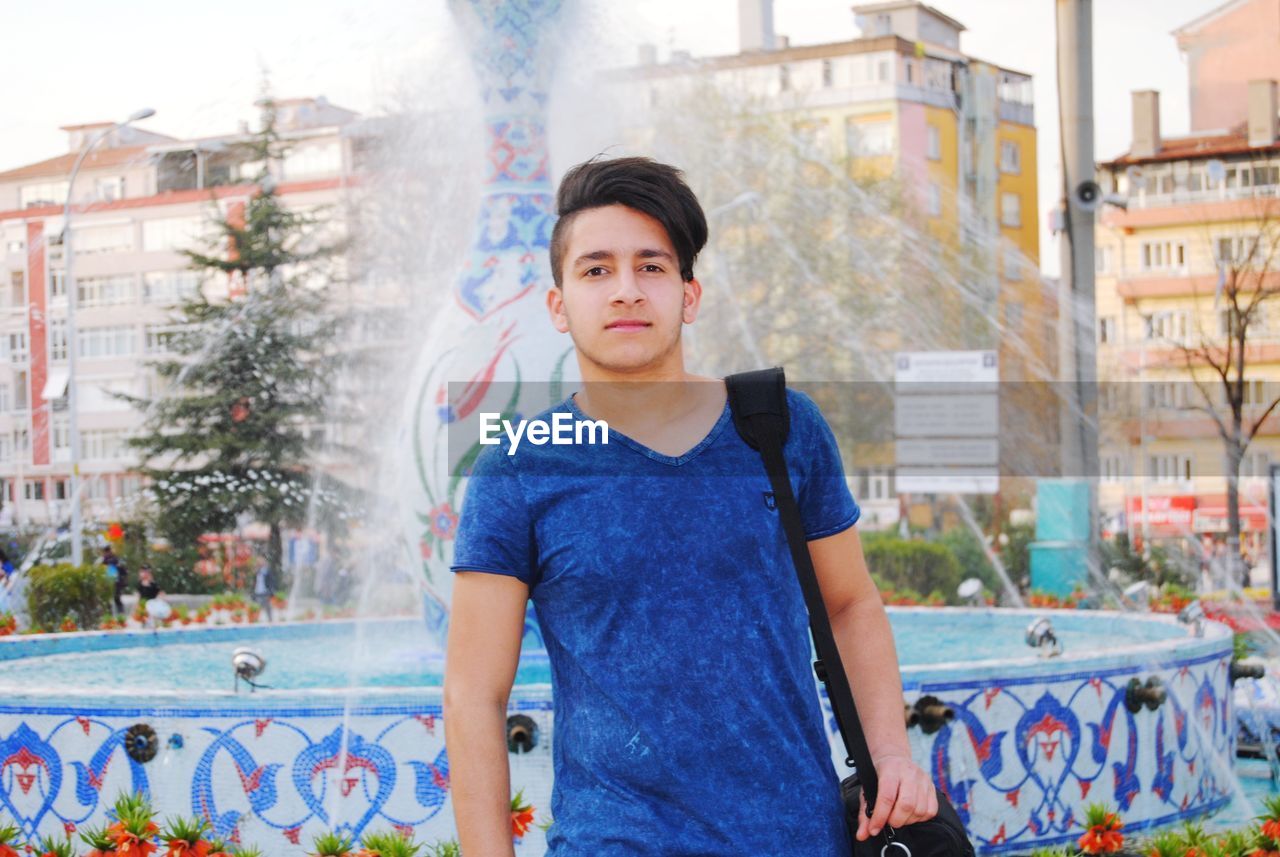 Portrait of young man standing against fountain in city