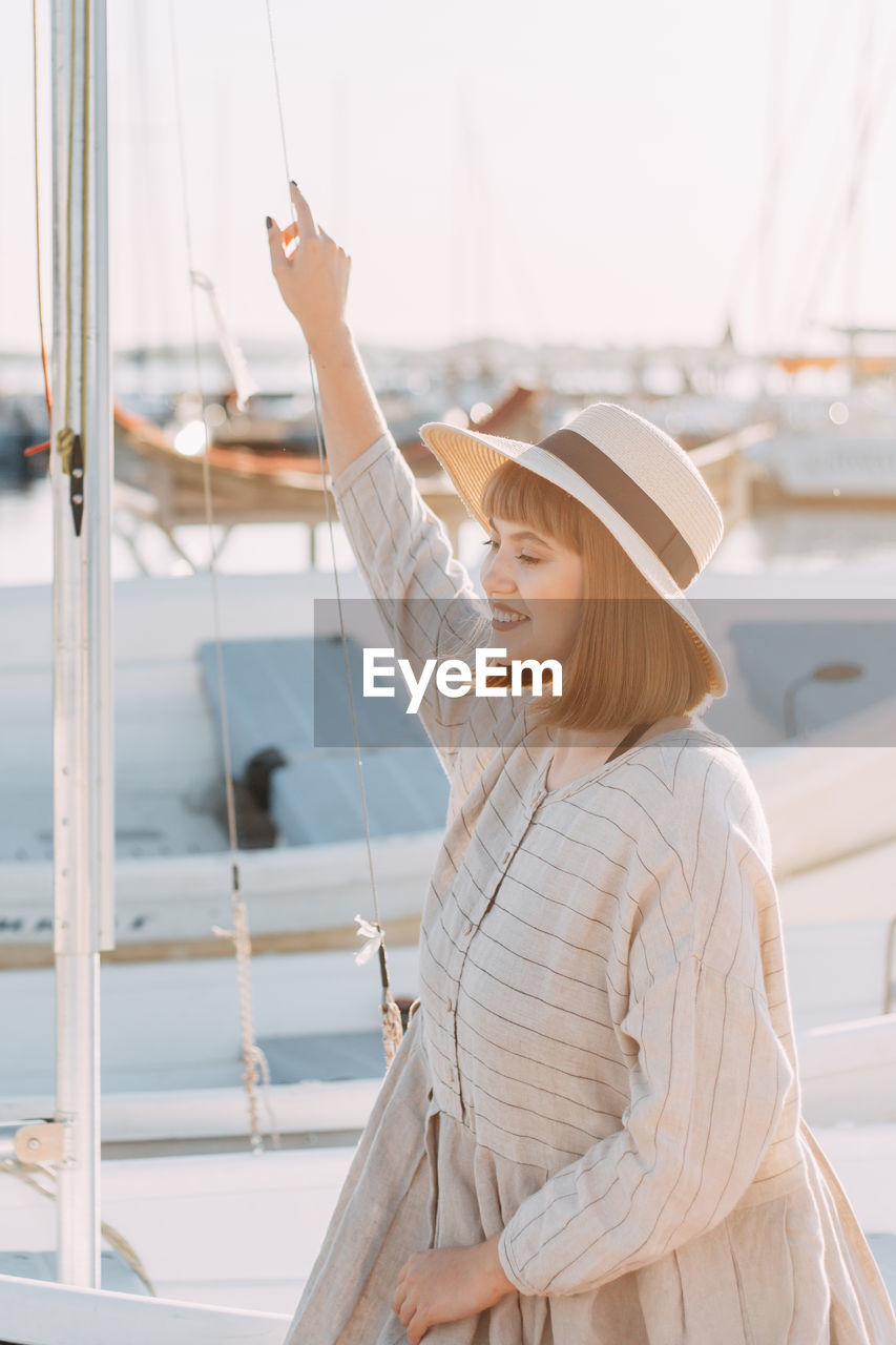 Happy young woman in dress and hat walks on the beach near the boats