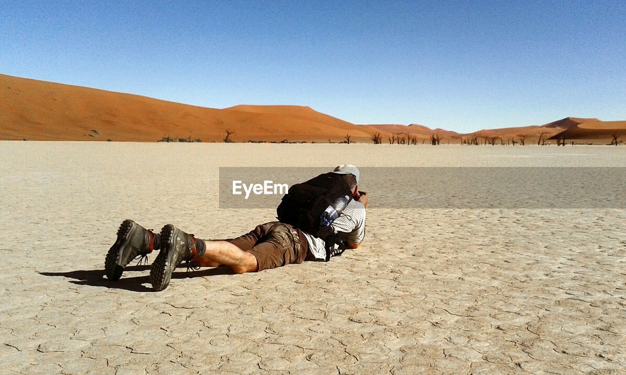 Full length of man lying on front at desert against clear blue sky