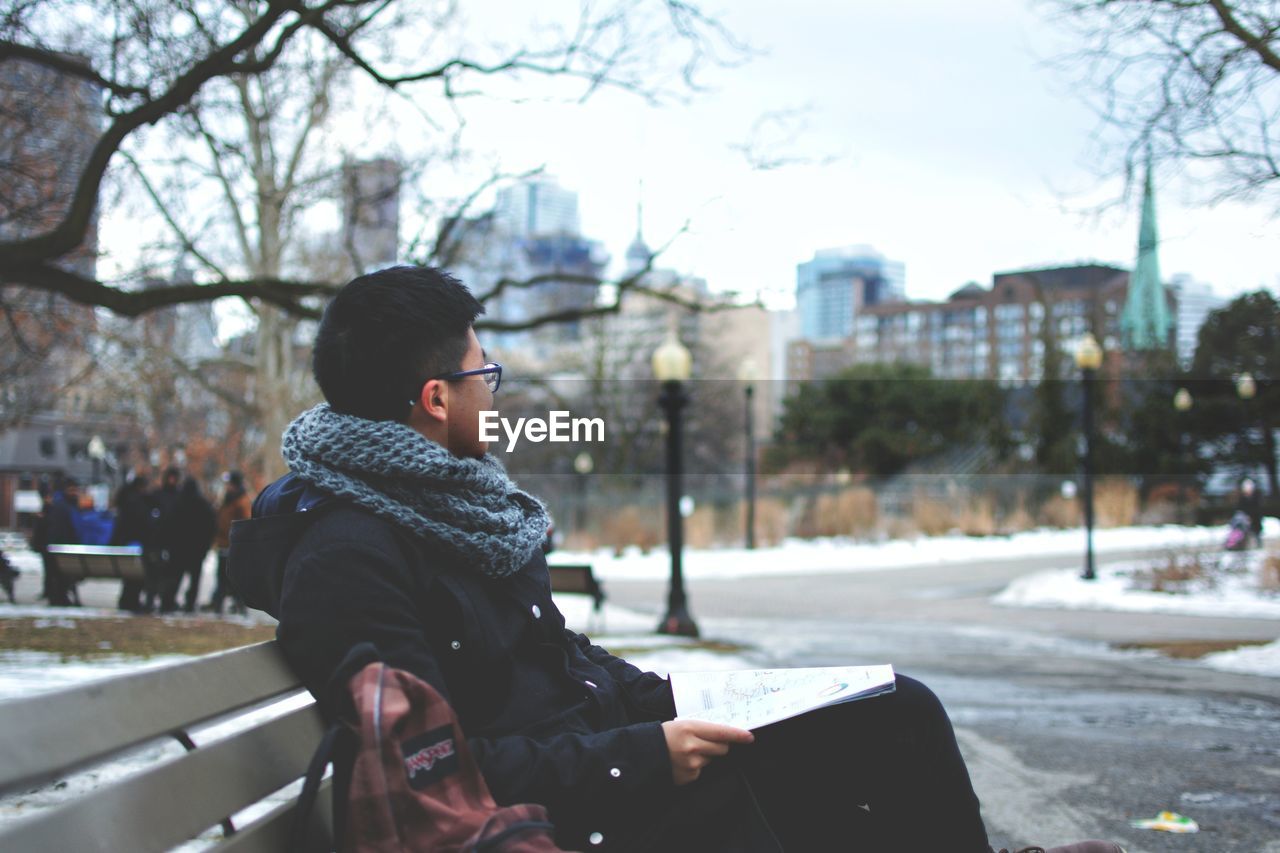 Side view of young man sitting on bench in city against sky