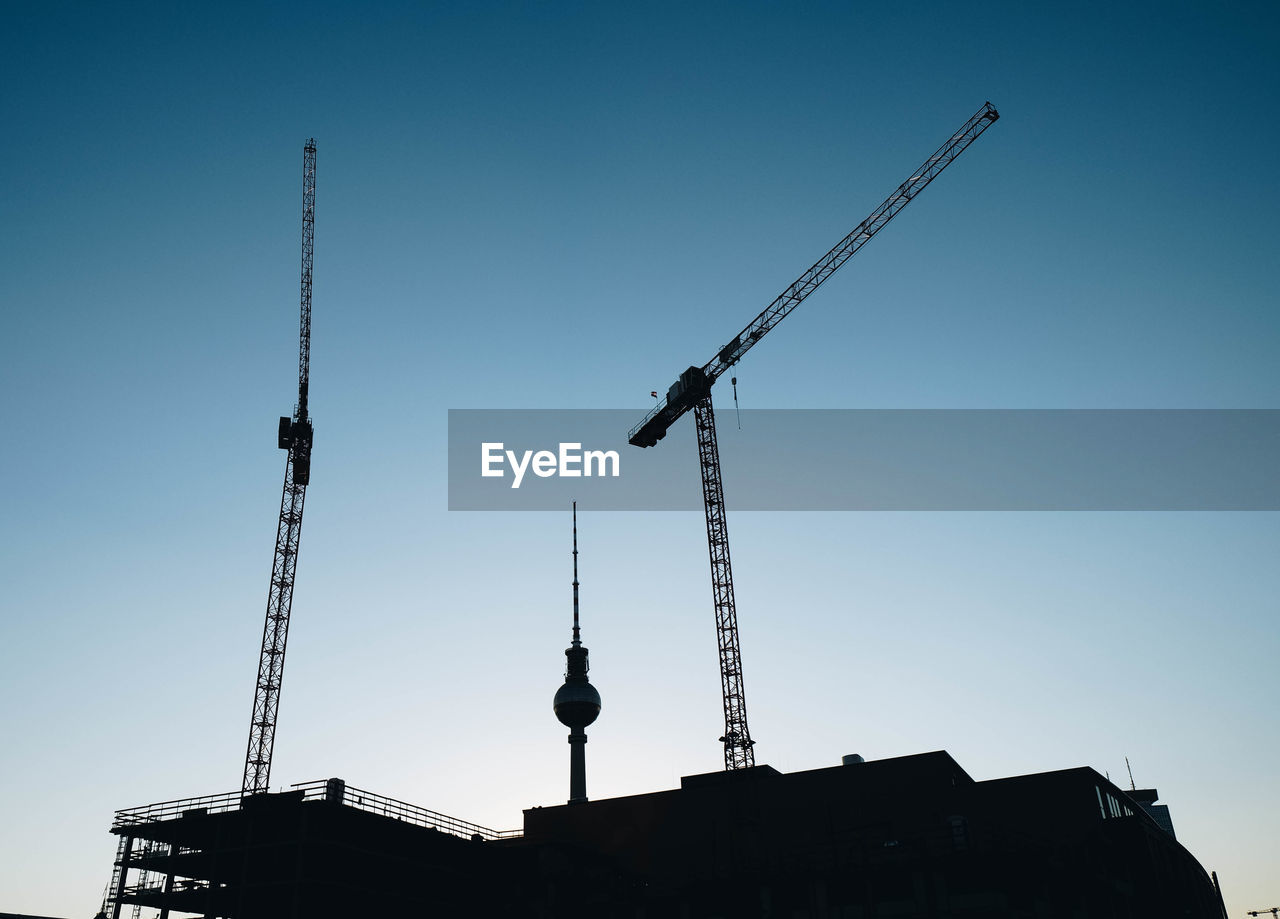 Low angle view of silhouette crane by building against clear blue sky