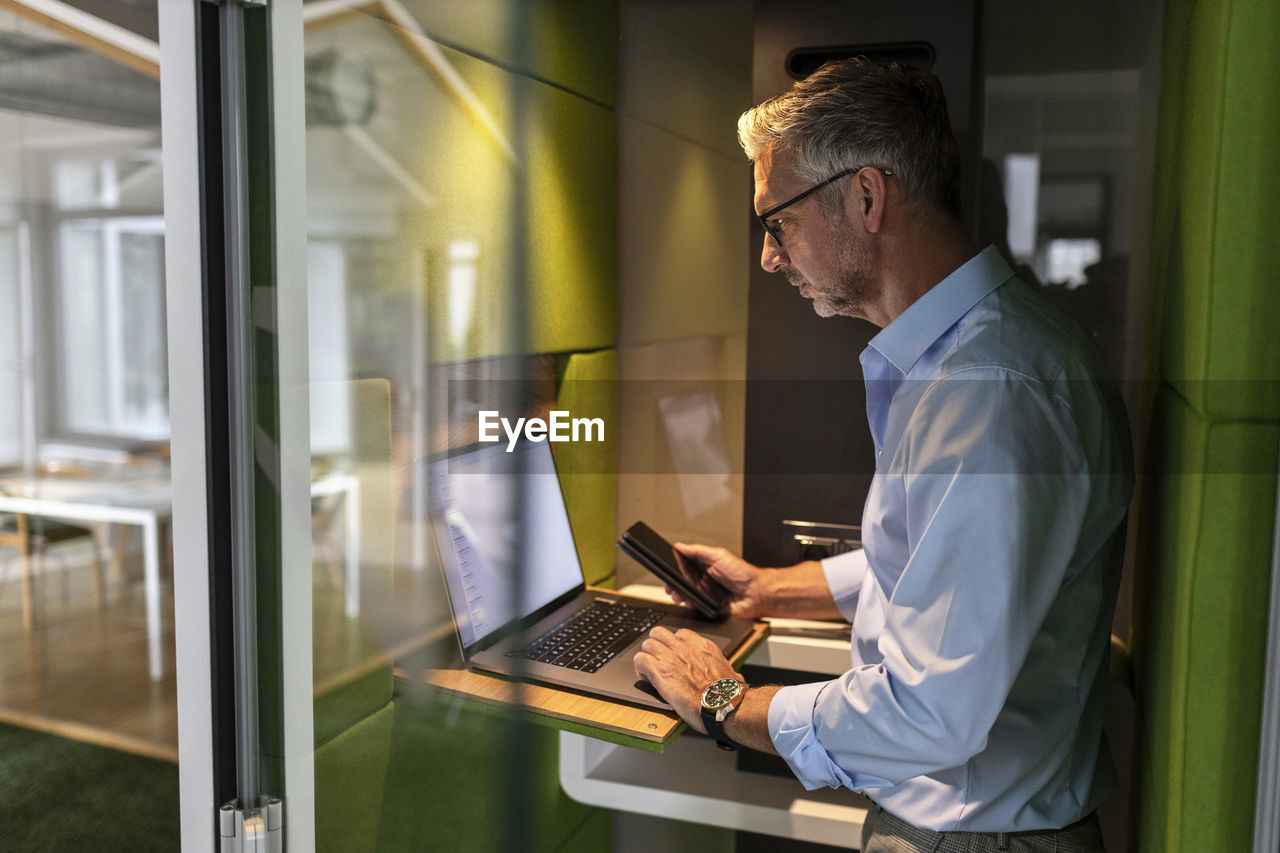Mature businessman holding mobile phone while using laptop in soundproof cabin at office