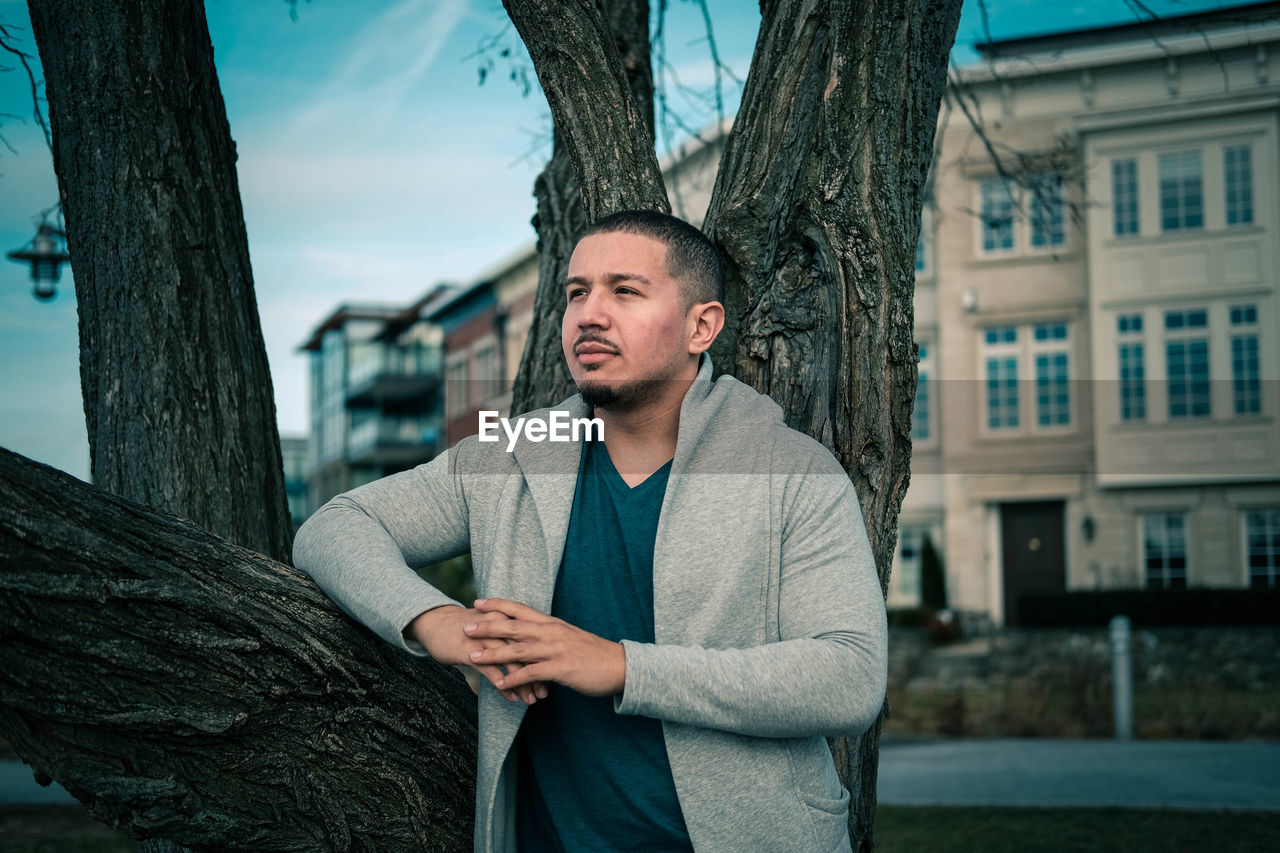 PORTRAIT OF YOUNG MAN STANDING AGAINST TREE TRUNK