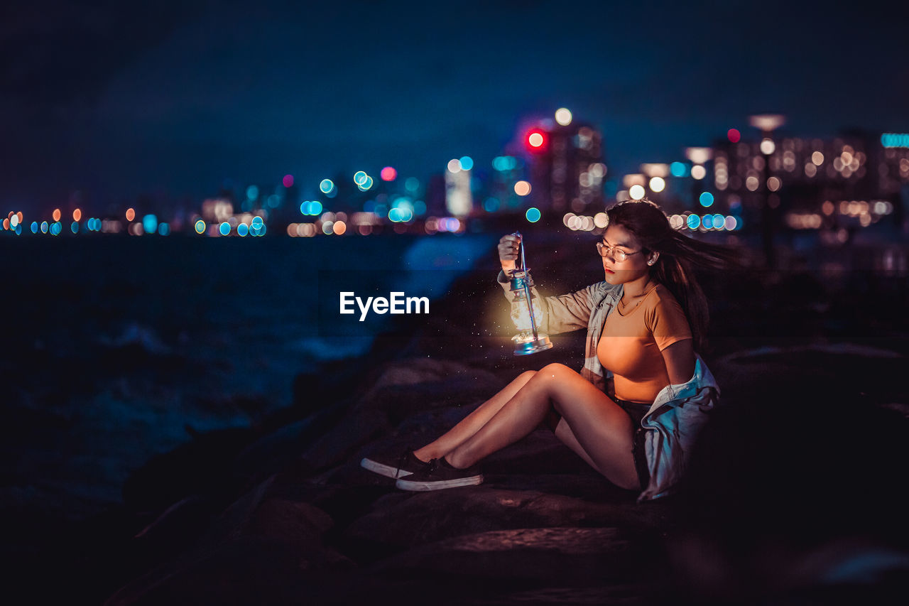 Woman sitting on thr sea wall holding a vintage lamp with city lights on the background.